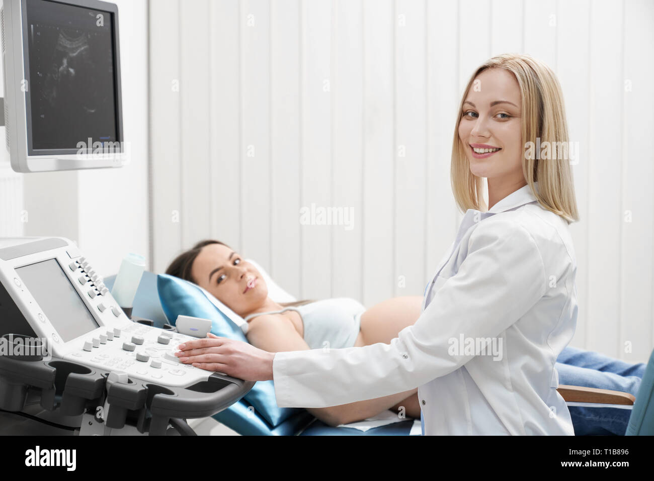 Schöne, hübsche Blondine Arzt tragen in Weiß Medical Gown, Ultraschall Untersuchung der Frau Bauch. Schwangere Frau liegend, mit Blick auf die Kamera lächelnd. Modernes Ultraschallgerät. Stockfoto