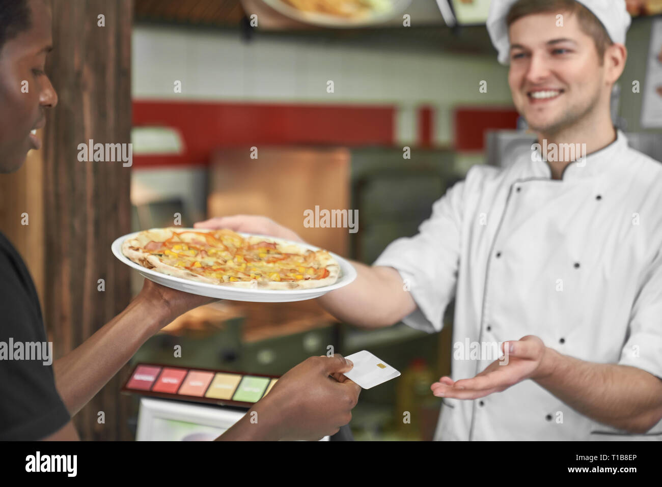 Junge afrikanische Holding leckere Pizza auf weiße Platte und das Bezahlen mit Karte. Schön, glücklich, Küchenchef in weiße Uniform Lächeln, das Gericht. Konzept des Fast Food und Pizzeria. Stockfoto