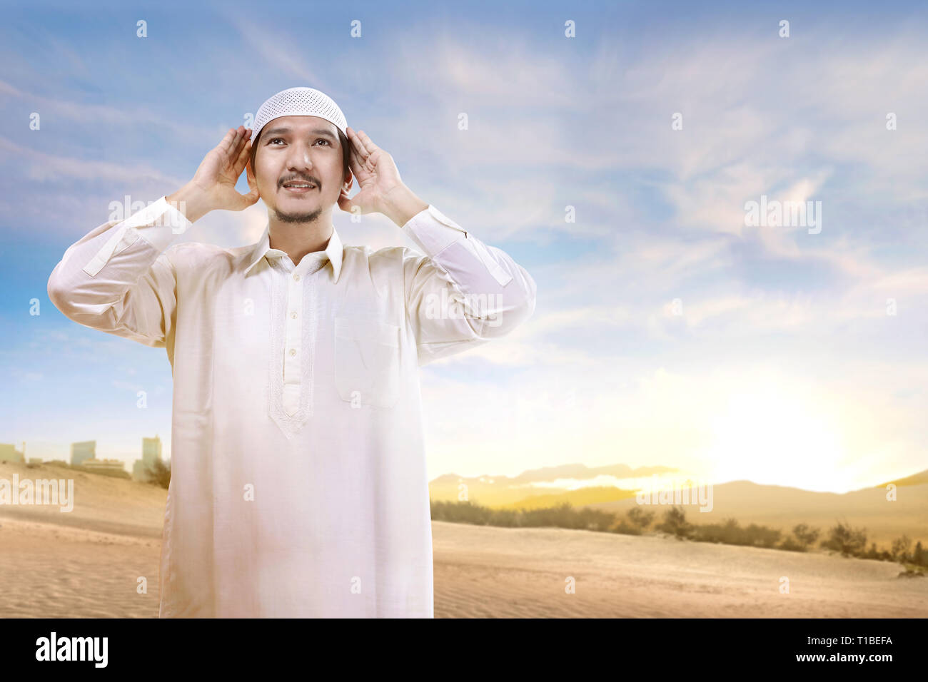 Lächelnd asiatischen muslimischen Mann mit Kappe stehen und beten in den Sand mit blauem Himmel Hintergrund Stockfoto