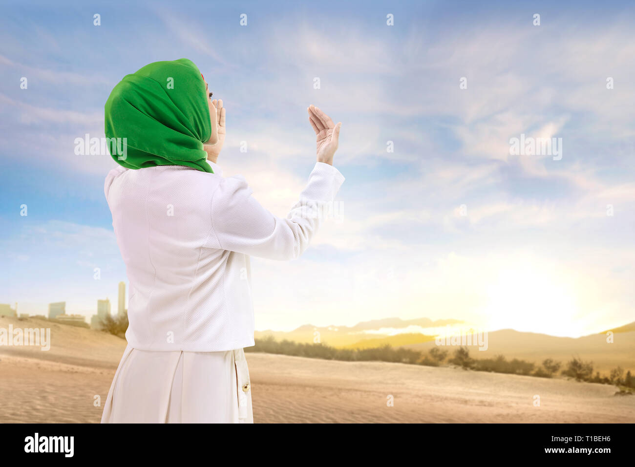 Schöne asiatische Muslimin in Schleier stehen und beten mit erhobenen Armen auf dem Sand mit blauem Himmel Hintergrund Stockfoto