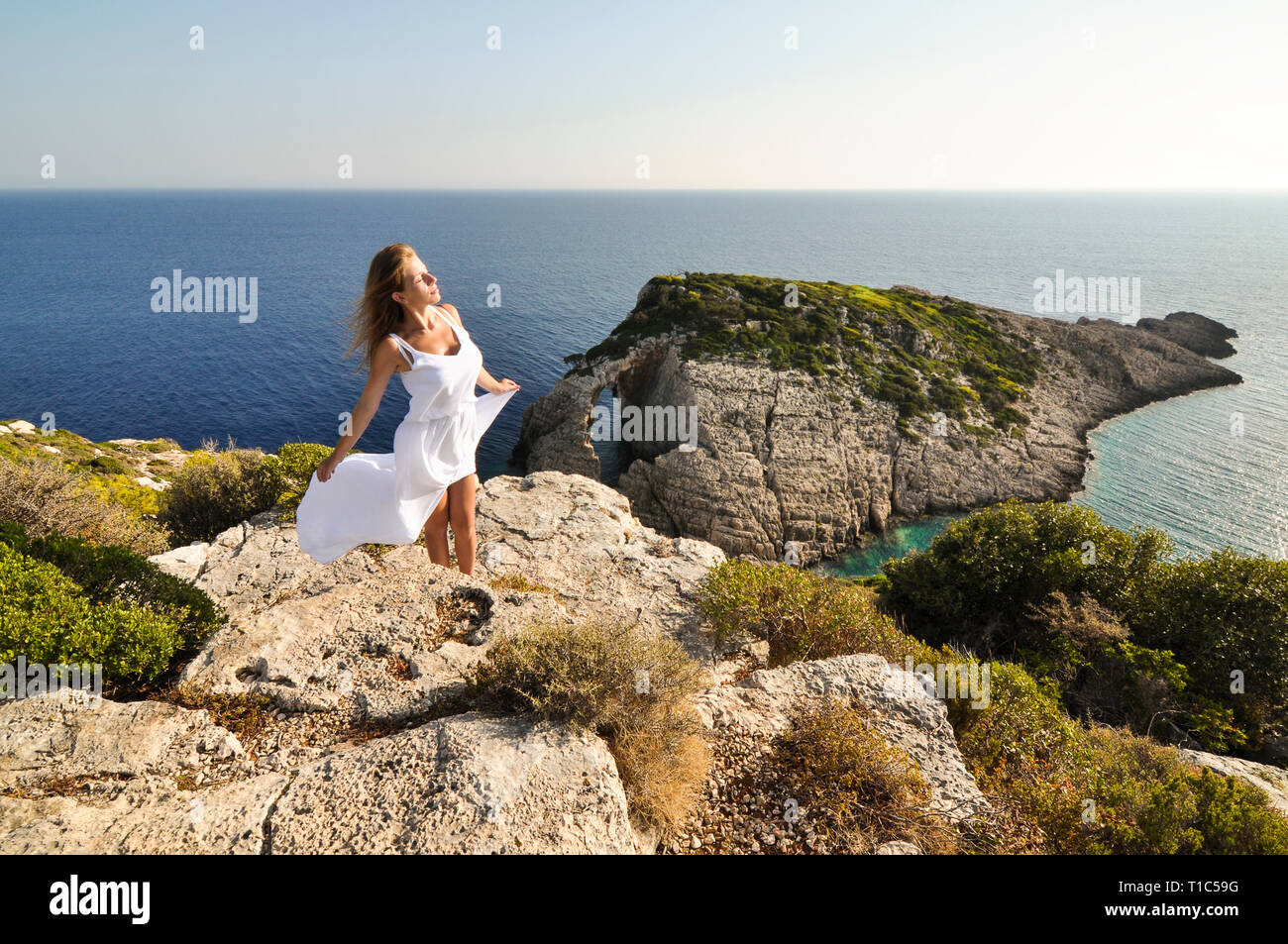 Frau in langen, weißen, winken Kleid steht auf der Klippe mit Blick von oben auf das Meer und die Insel. Sonniger Meereslandschaft mit weiblichen Touristen fühlen sich du Stockfoto