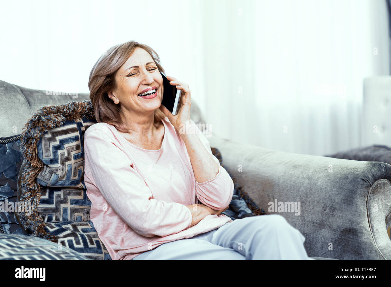 Ältere Frau mit einem Telefon in der Hand in guter Stimmung ruht sich zu Hause im Wohnzimmer. Stockfoto