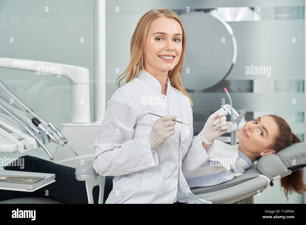 Charmante weibliche Zahnarzt posieren. Schöne Frau lächelnd, Kamera und Holding Restaurierung Instrumente. Client hinter Lügen auf Zahnheilkunde Stuhl. Gerne Arzt im weißen Kittel tragen. Stockfoto
