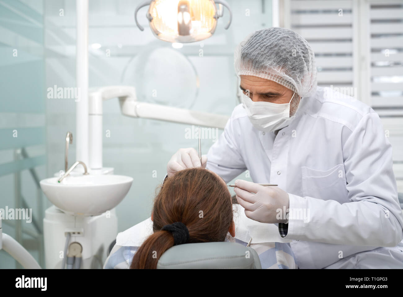 Zahnarzt Behandlung der Zähne des Client in private Zahnheilkunde Klinik. Stomatologist tragen im weißen Kittel, medizinische Maske, Mütze und Handschuhe. Lampe, weiß Waschbecken und zahnmedizinischen Ausrüstung im Schrank. Stockfoto