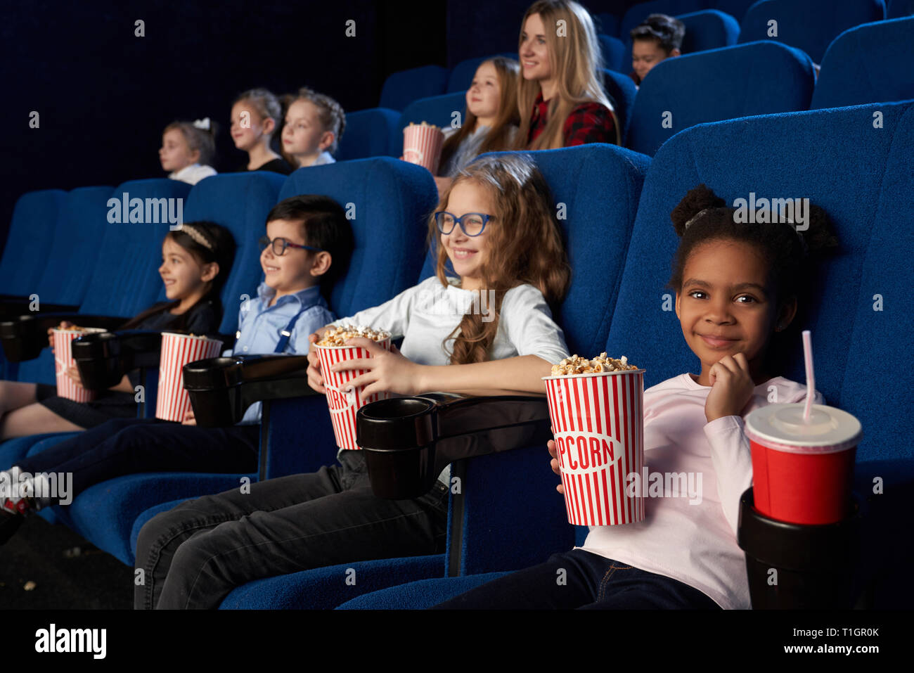 Gruppe von Kindern im Kino sitzen, sich Zeichentrickfilme, genießen Premiere. Nette, hübsche Afrikanische Mädchen, dass Popcornwanne, an der Kamera suchen, lächelnd. Stockfoto