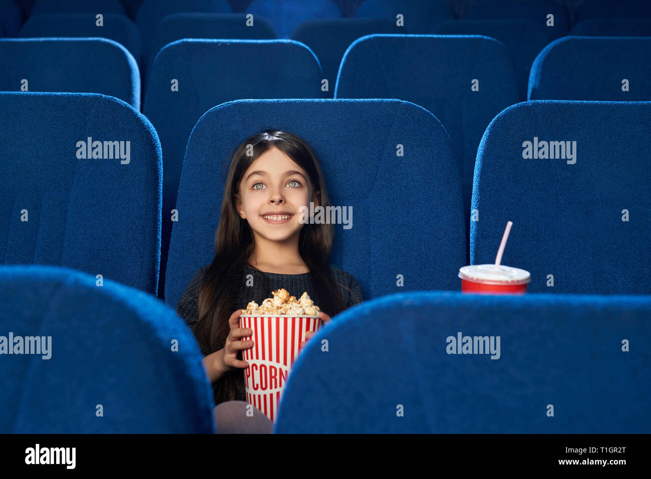 Schön, wunderschöne kleine Mädchen allein sitzen im Kino, in komfortablen Dunkelblau Stuhl. Glückliches Kind lächelnd, halten große Popcornwanne, aufzupassen Film. Stockfoto