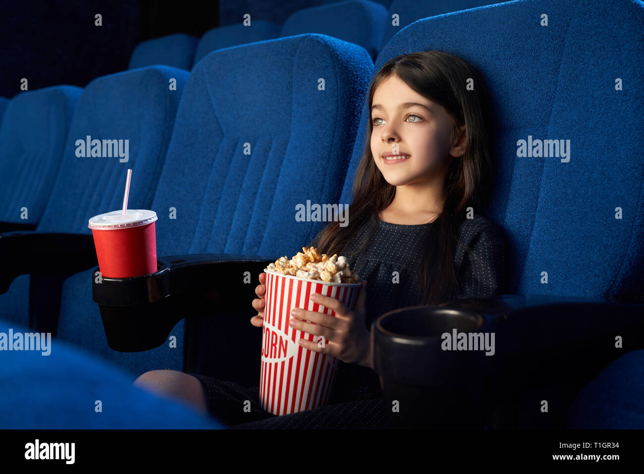 Niedlich, hübsch, schöne Mädchen holding Popcornwanne, sitzen in bequemen Stuhl im Kino. Fröhliches Kind beobachten, genießen Film oder Cartoon. Stockfoto