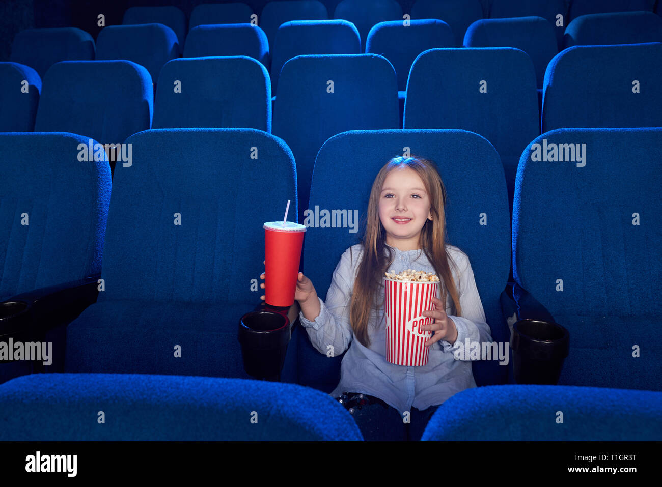 Vorderansicht des schönen Kind lächelnd, posierend, an der Kamera schaut. Glückliche, fröhliche Mädchen im Kino sitzen, holding Popcornwanne, rot Pappbecher mit Kohlensäure trinken. Stockfoto