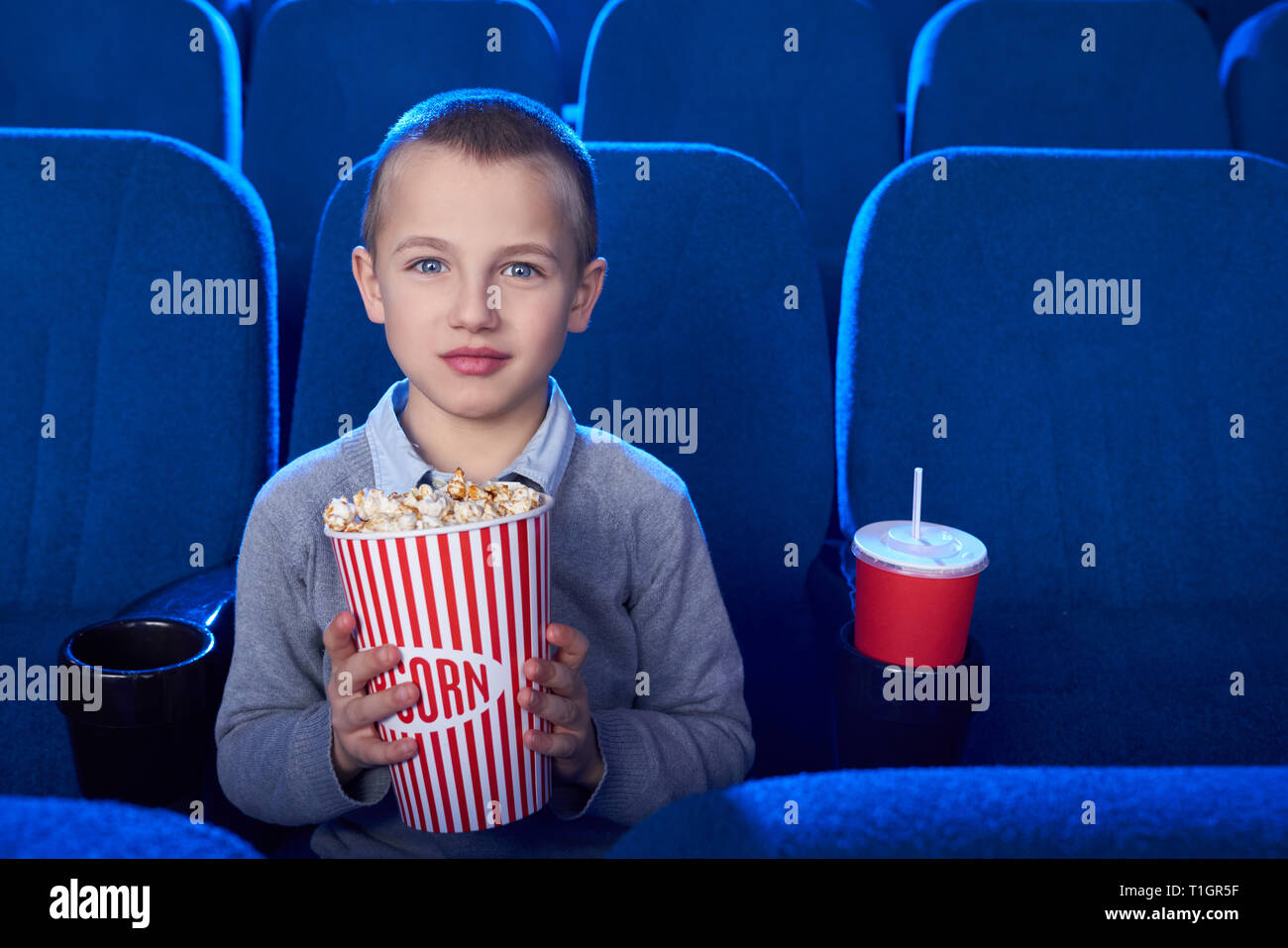 Vorderansicht des hübschen kleinen Jungen im bequemen Sessel sitzen im Kino, auf Kamera posieren. Fröhliches Kind holding Popcornwanne. Stockfoto