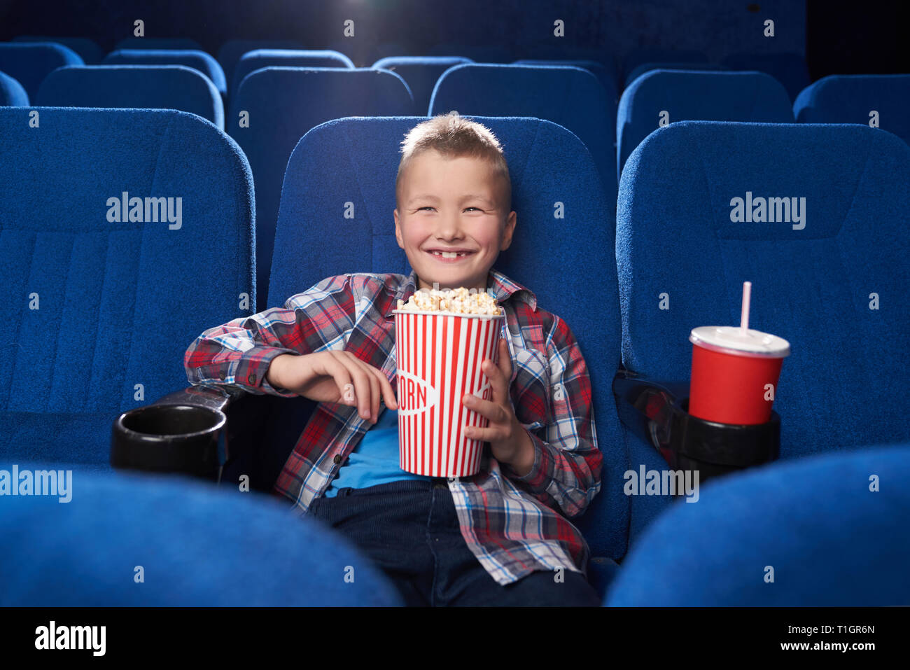 Lustige, fröhliche Zicklein aufzupassen Film oder Cartoon Komödie im Kino. Fröhlicher Junge, Lächeln, Lachen, halten große Popcornwanne, sitzen in Dunkelblau bequemen Stühlen. Stockfoto