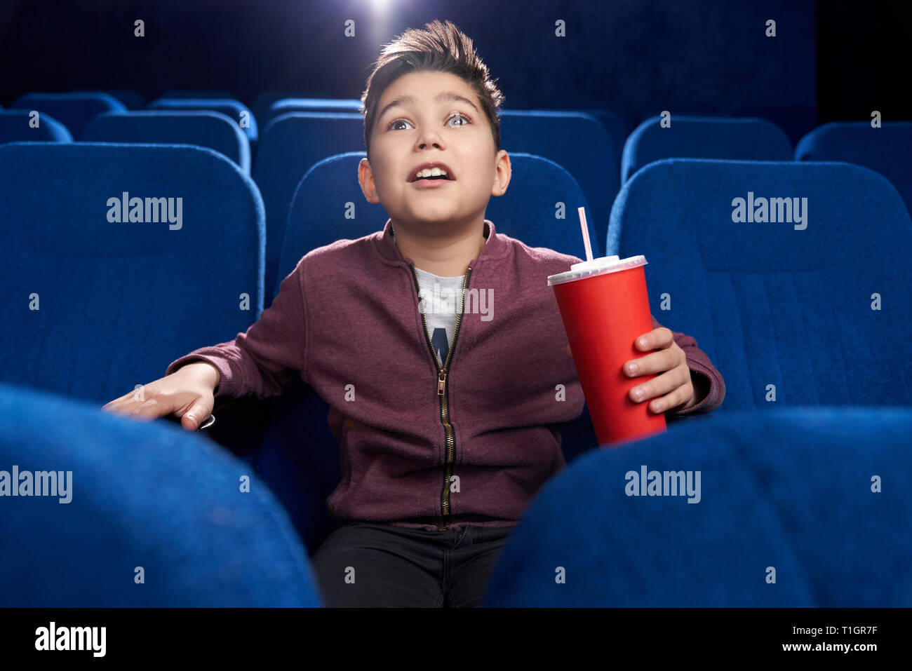 Schockiert, fasziniert Junge im Kino sitzen, am Bildschirm aufmerksam schauen mit geöffnetem Mund. Teenager halten roten Pappbecher mit Kohlensäure trinken. Stockfoto