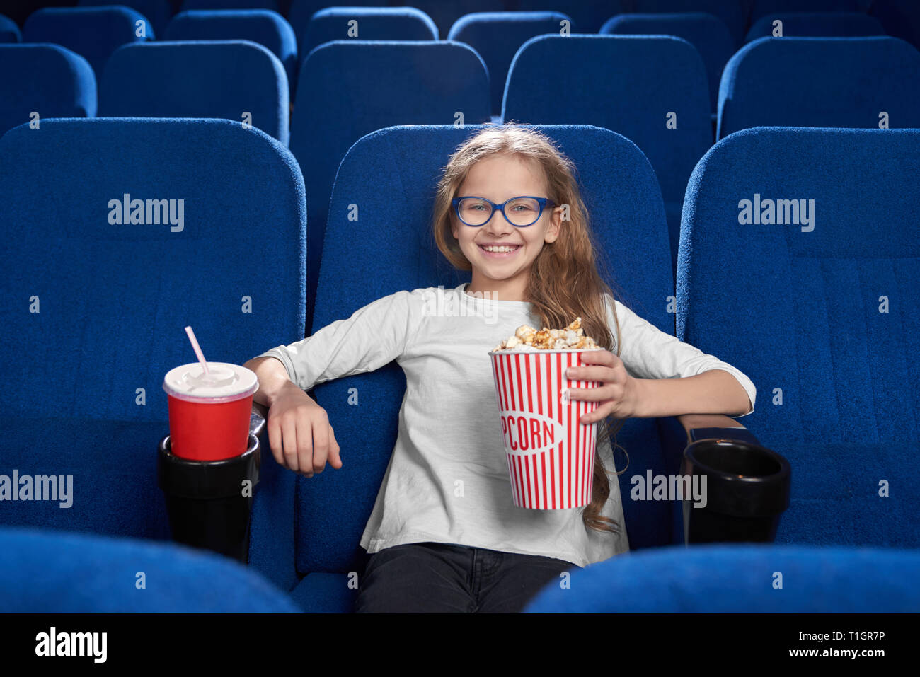 Vorderansicht des Glücklichen, freudigen Mädchen im Kino sitzen, holding Popcornwanne, rot Pappbecher mit Kohlensäure trinken. Schöne Kind lächelnd, posierend, an der Kamera schaut. Stockfoto