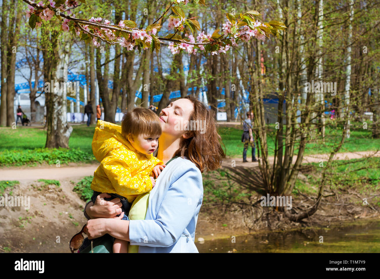 Junge Frau mit einem Kind auf dem Arm zufrieden Sakura Blüten im Frühling Park. Freude an der Mutterschaft und die Entwicklung des Kindes. Kirschblüte Baum im späten Frühjahr. Flora im Nordwesten von Russland, St. Petersburg Stockfoto