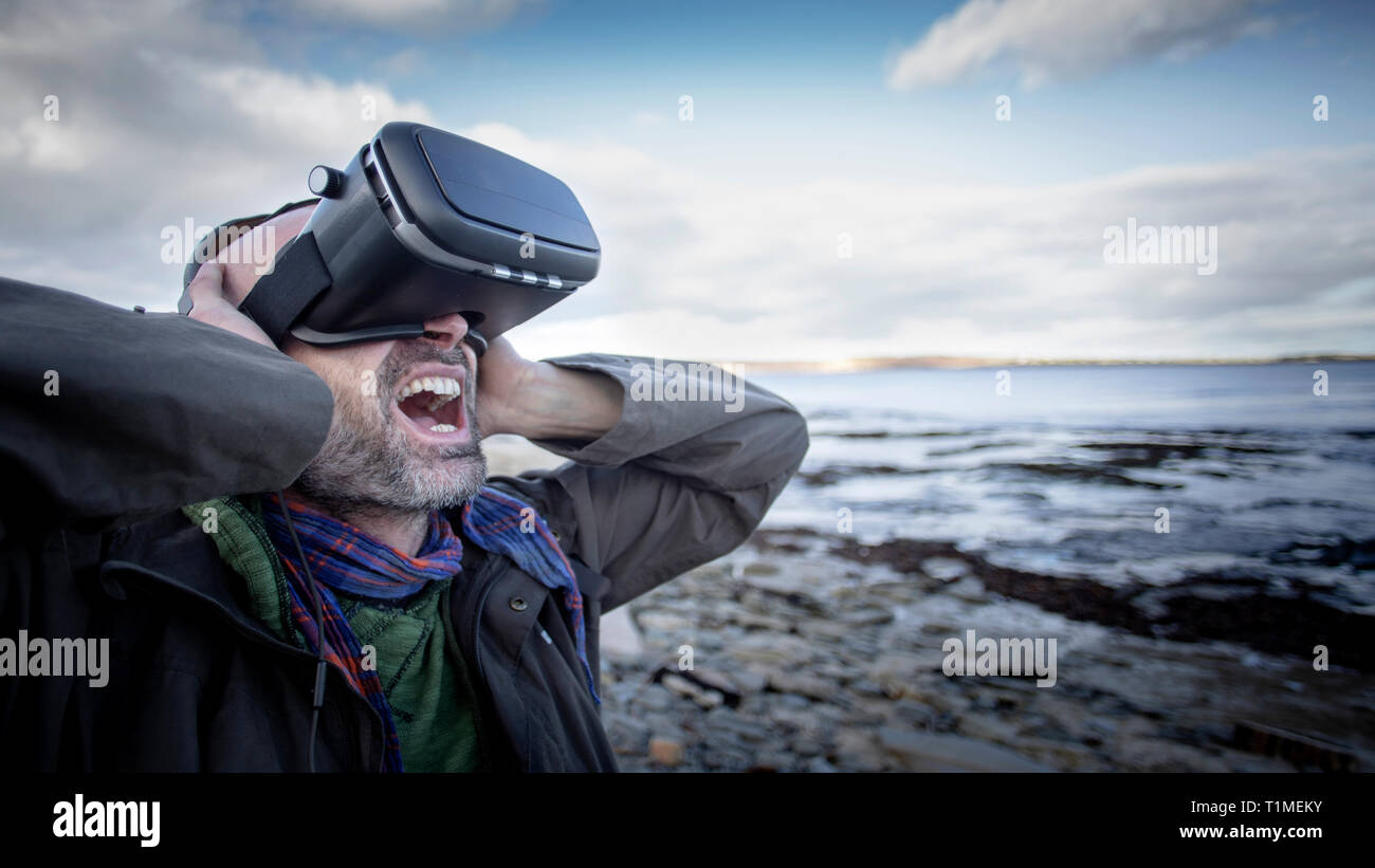 Eine Frontansicht Schuß eines reifen kaukasischen Mann mit einem Virtual reality Headset während an einem Strand im Vereinigten Königreich stand, hat er seine Arme angehoben Stockfoto