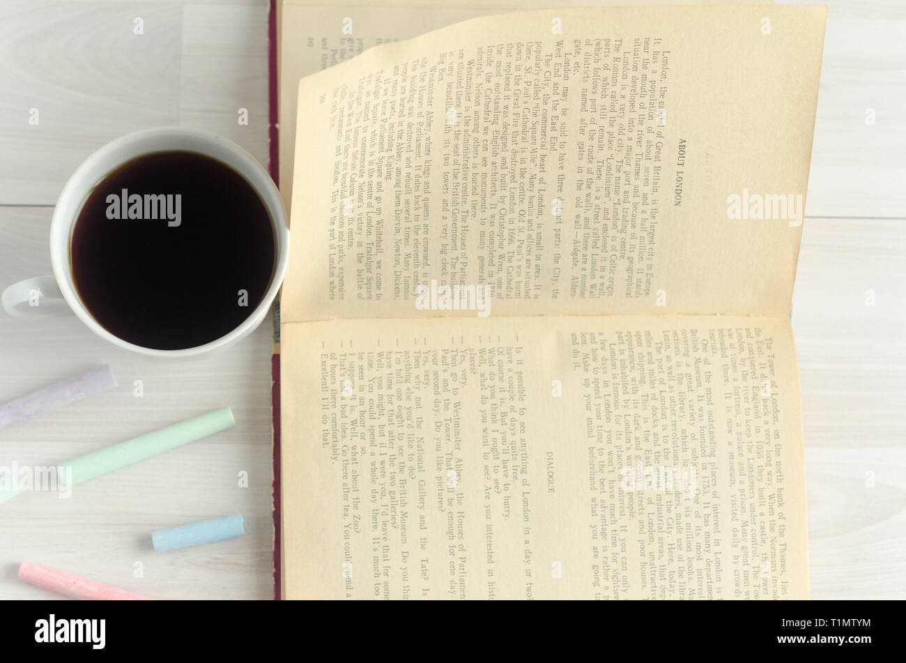 Eine Tasse Kaffee, bunte Kreide und ein offenes Buch auf weißem Holz- Hintergrund. Welttag des Buches, flach. Stockfoto