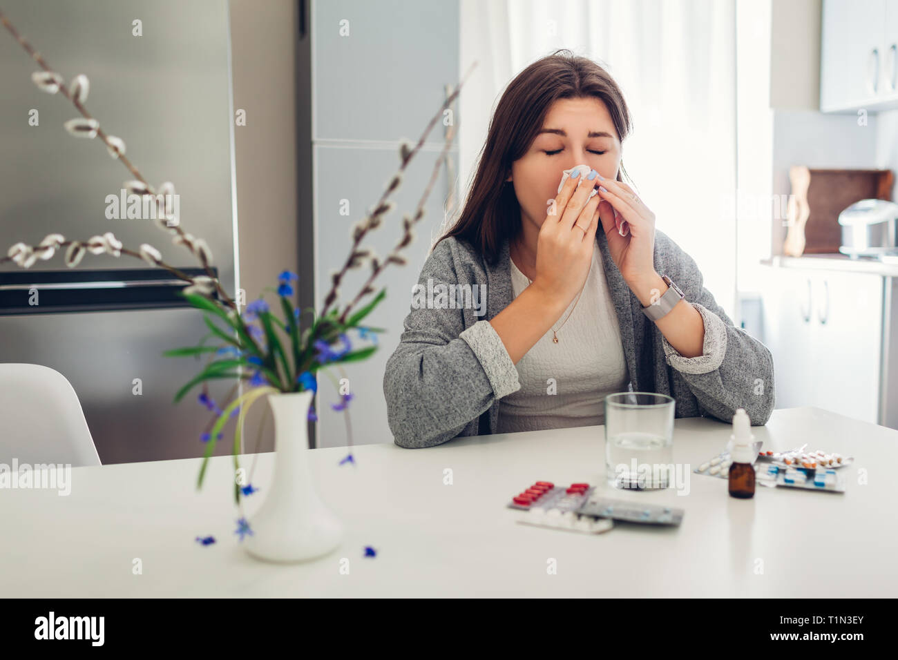Frühling Allergie. Junge Frau Niesen, weil der Blumen mit Pillen auf Küche zu Hause umgeben. Saisonale Allergien Konzept. Stockfoto