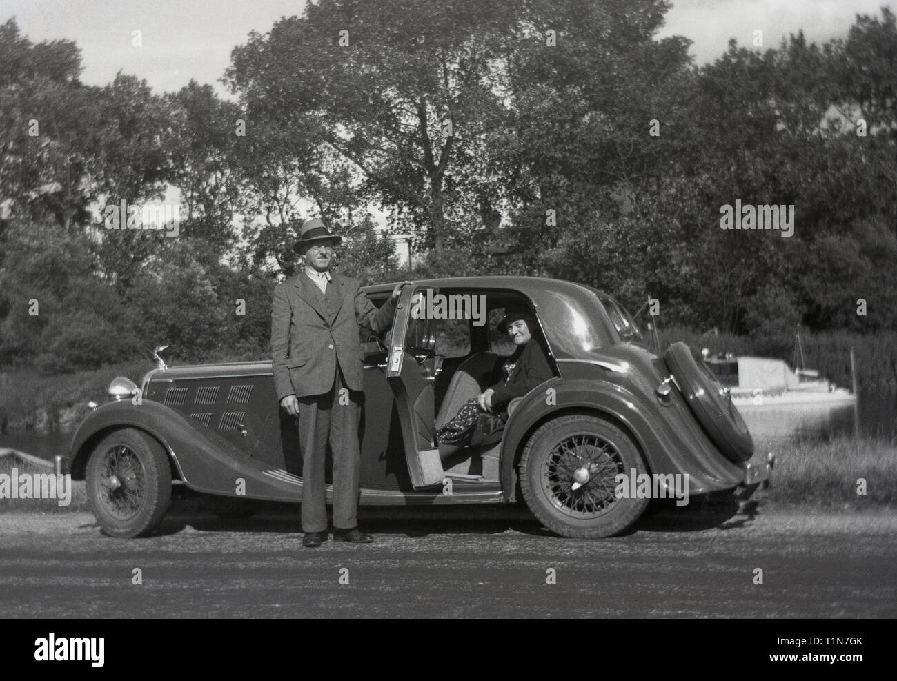 1930er Jahre, historische, Autofahren in dieser Ära, ein Herr im Anzug und einen Hut tragen, öffnet die Hintertür von seinen Sport Auto für seine weiblichen Passagier, wie sie für eine Pause am Ufer eines Flusses, England, UK. Stockfoto