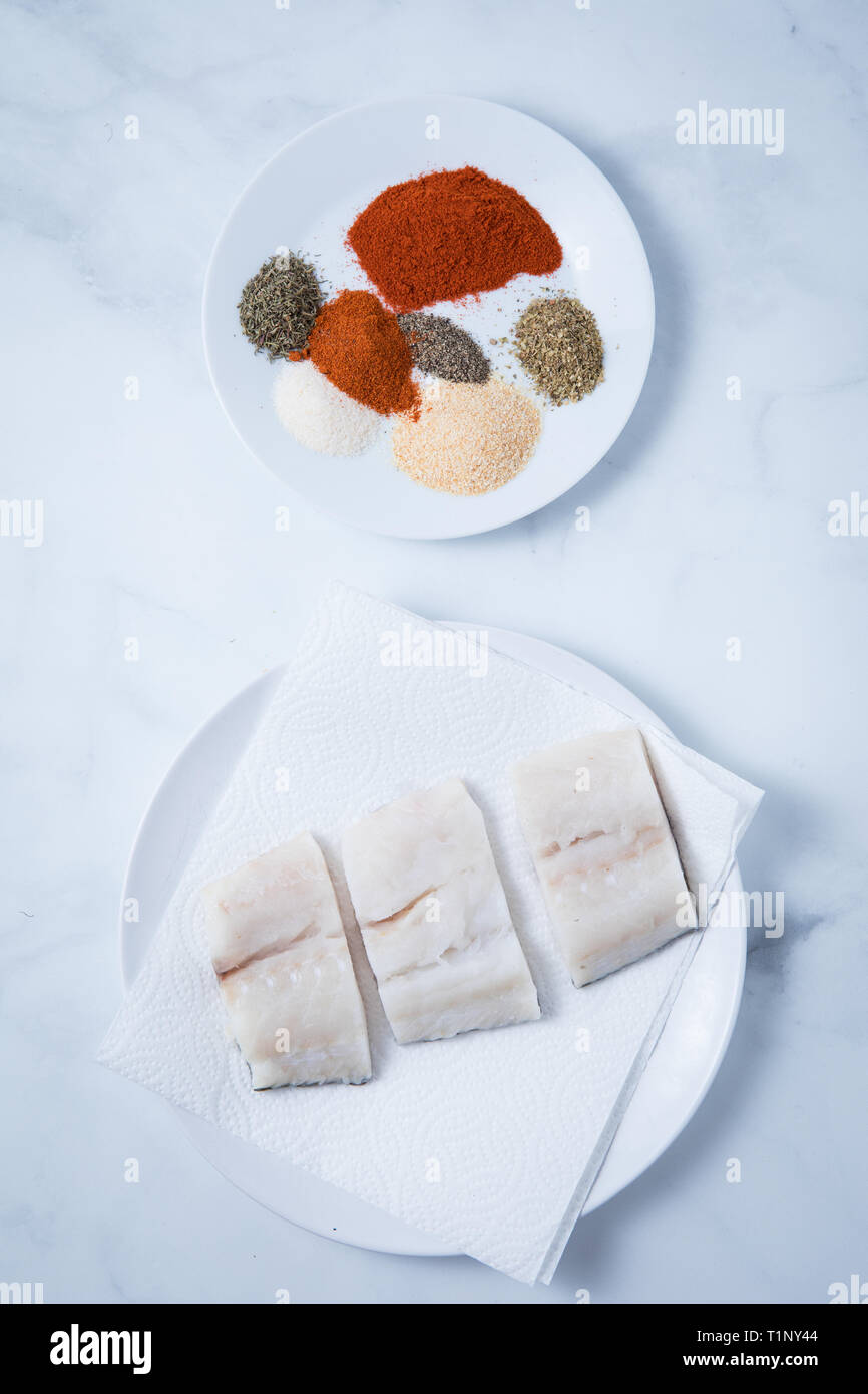 Raw Kabeljau Fischfilets auf Servietten und eine runde weiße Platte mit Gewürze auf einem weißen Hintergrund. Rohe Stücke von cod. Stockfoto