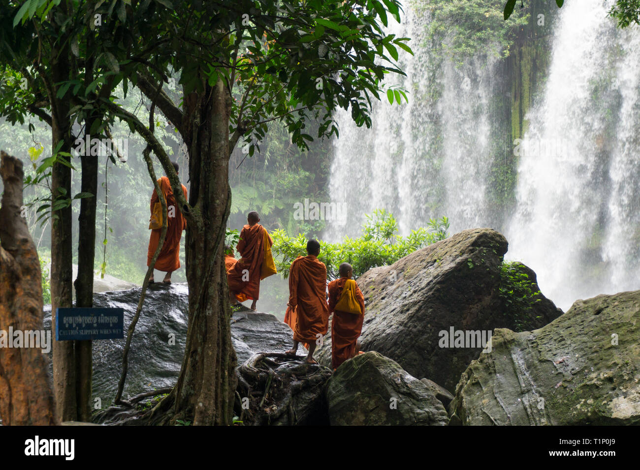 Eine Gruppe junger buddhistischer Mönche tragen die traditionelle orange Kleider stehen auf große Felsbrocken und genießen den Blick auf den Wasserfall Stockfoto