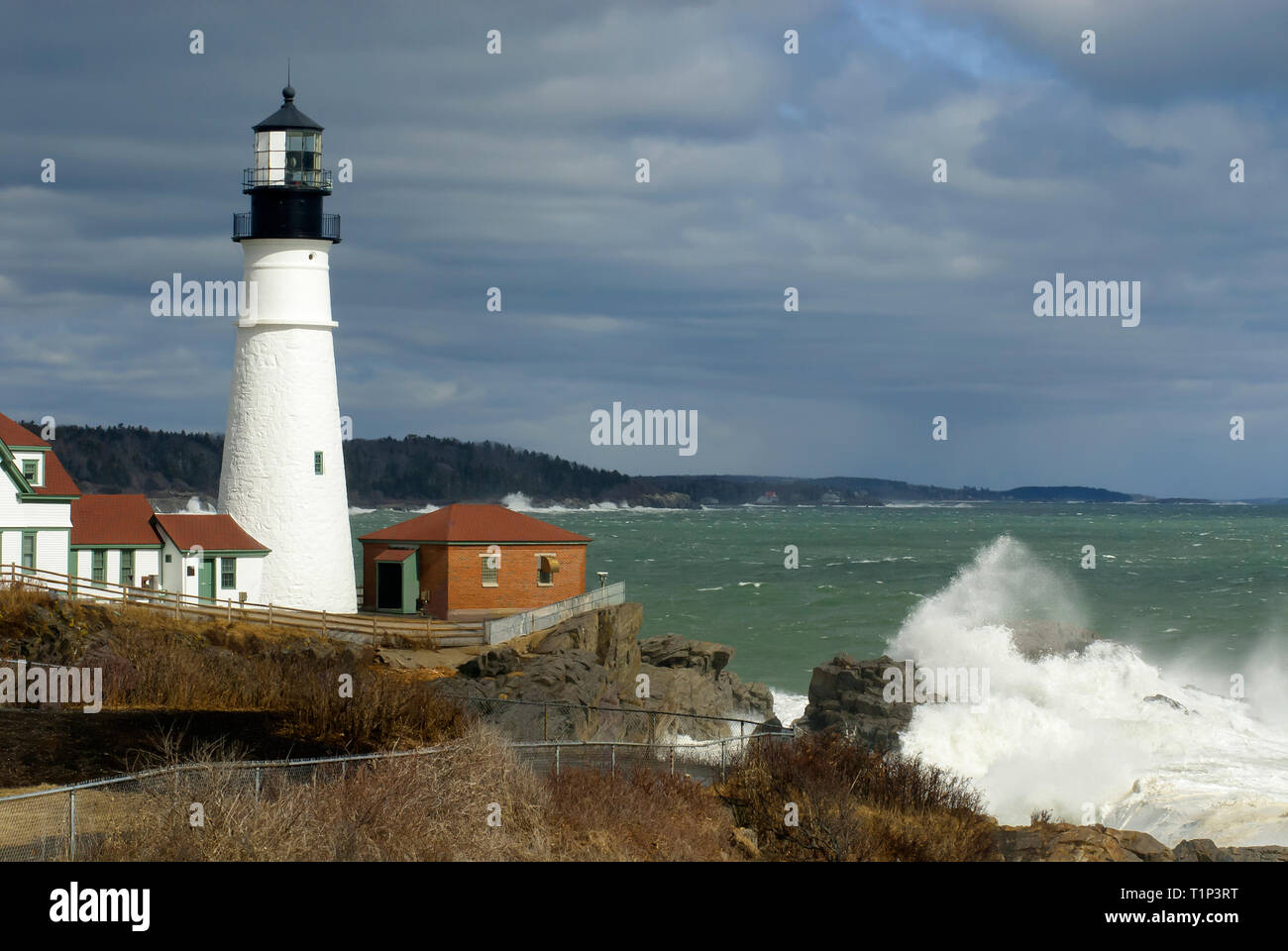 Sonnenlicht beleuchtet Portland Head Lighthouse, durch Gewitterwolken, wie riesige Welle bricht über Felsen, in Maine. Stockfoto