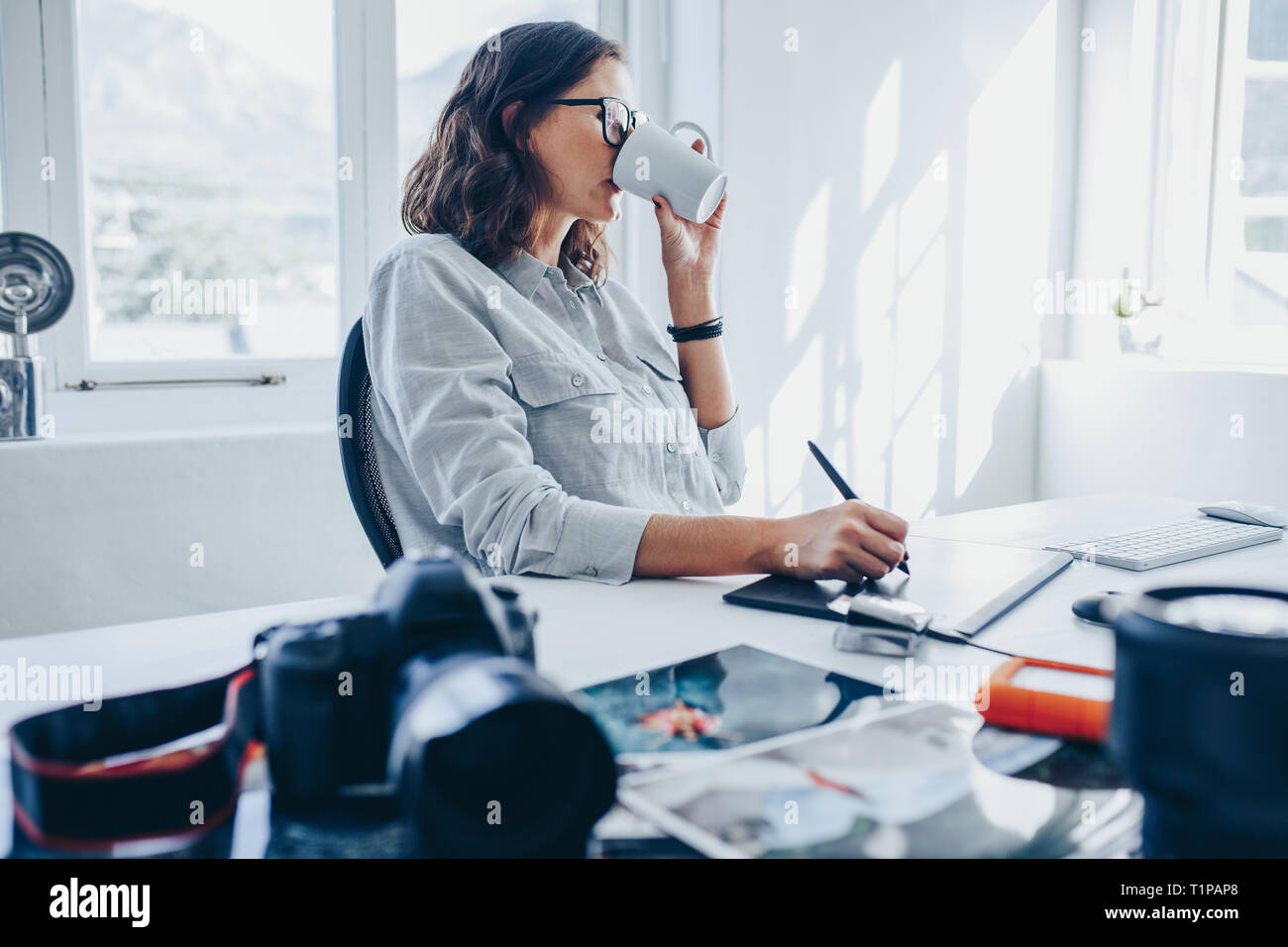 Frau artist Kaffee trinken während der Arbeit am Schreibtisch. Fotografin Retuschieren von Fotos in Büro- und trinken eine Tasse Kaffee. Stockfoto