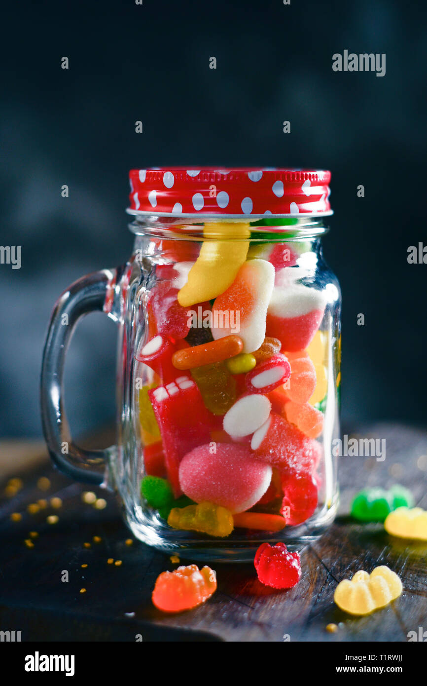 Ein Glas Marmeladenglas voller Bonbons mit verschiedenen Aromen und Farben, auf einem rustikalen Holztisch, gegen einen dunklen Hintergrund Stockfoto
