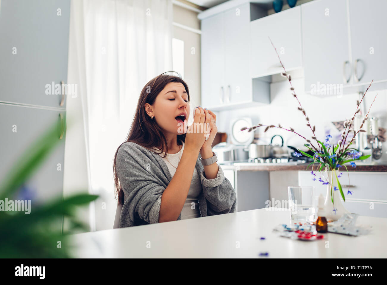 Frühling Allergie. Junge Frau Niesen, weil der Blumen mit Pillen auf Küche zu Hause umgeben. Saisonale Allergien Konzept. Stockfoto