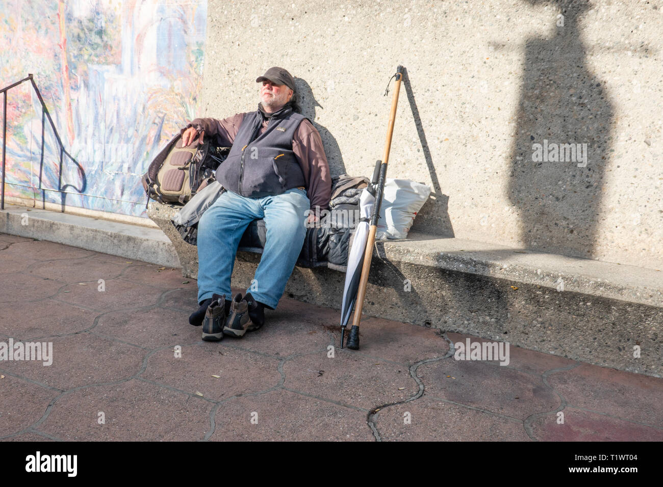 Ein obdachloser Mann sitzt mit seinen Schuhen weg auf eine Stadt konkrete Bank mit seinen Spazierstock, Regenschirm und Rucksack neben ihm auf der State Street in Santa Ba Stockfoto