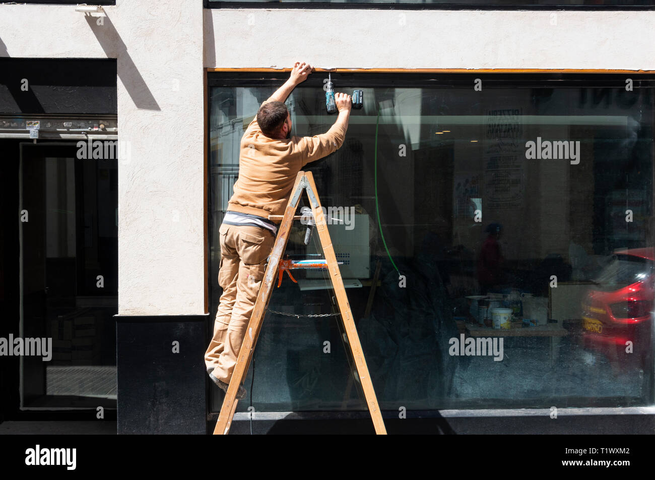 Arbeiter auf einer Leiter, der eine Reparatur mit einer Akku-Bohrmaschine in Sevilla, Spanien, macht Stockfoto