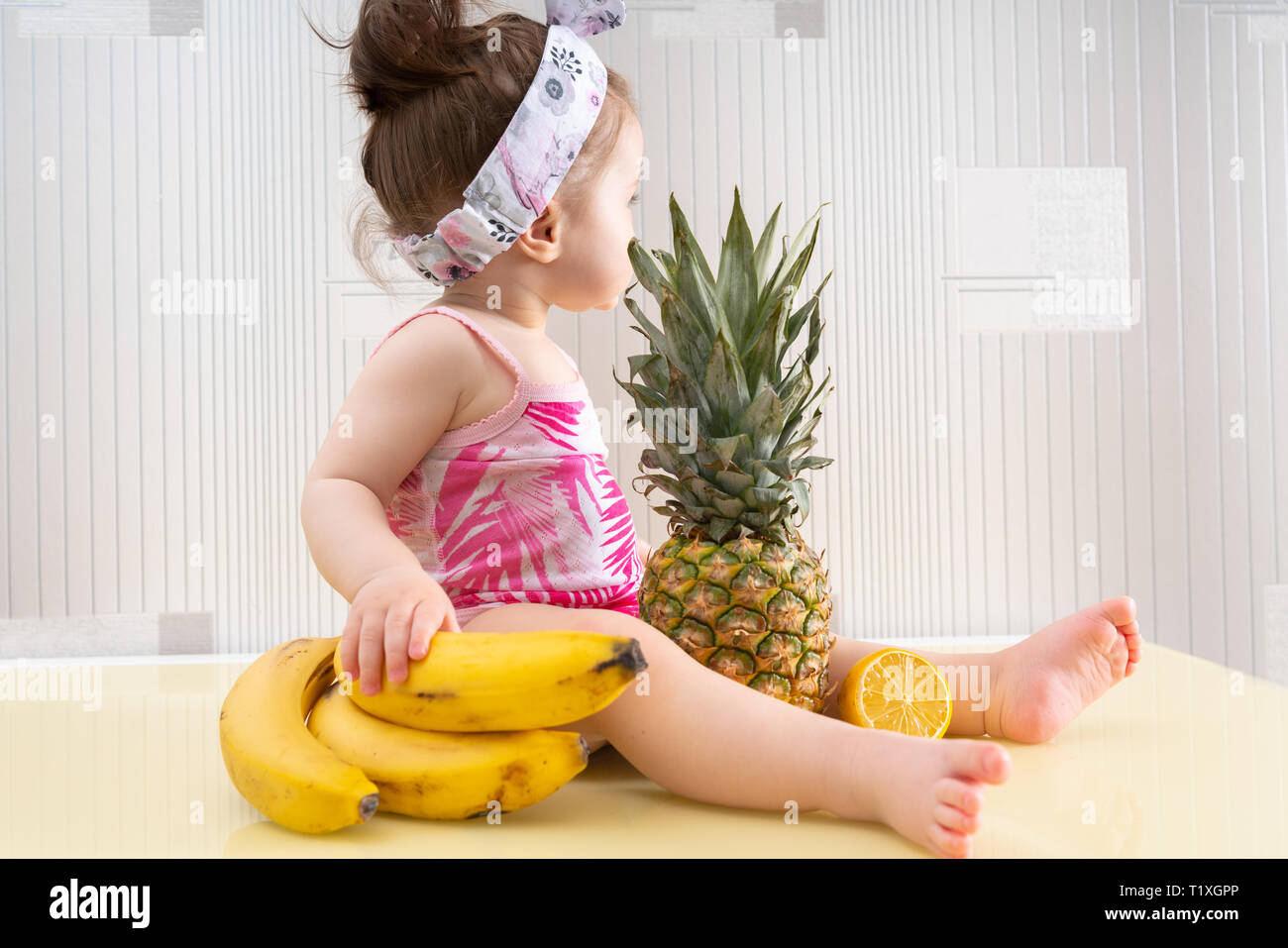 Kleine Mädchen in rosa Hemd und weißem Haar Gurt sitzt am Tisch mit tropischen Früchten und Wegsehen. Große reife Ananas vor ihr, Zitrone Stockfoto