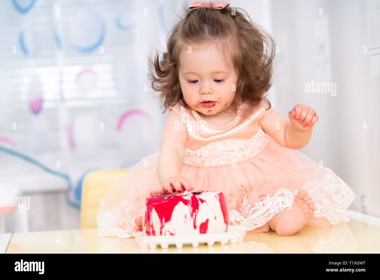 Unordentliche hübsches kleines Mädchen in ein rosa Kleid essen eine Geburtstagstorte mit Marmelade bestrichenen Rund um den Mund und klebrige Hände, als sie zusammen auf einem Tisch sitzt Stockfoto