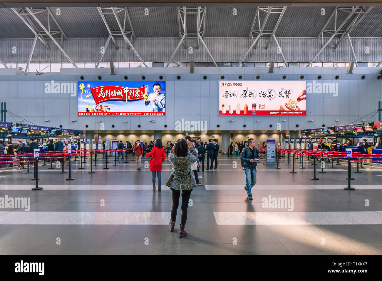 Beijing Capital Airport T2 Interieur mit Reklametafeln, Zähler und Reisende. Frau ist Fotografieren mit Smart Phone. Stockfoto