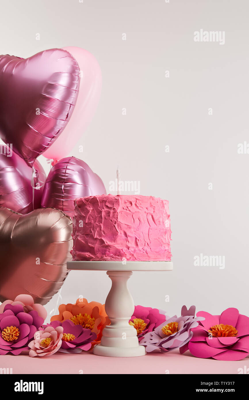 Rosa Geburtstagskuchen mit Kerze für Kuchen stehen in der Nähe von Papier Blüten und herzförmigen Luftballons auf Grau Stockfoto