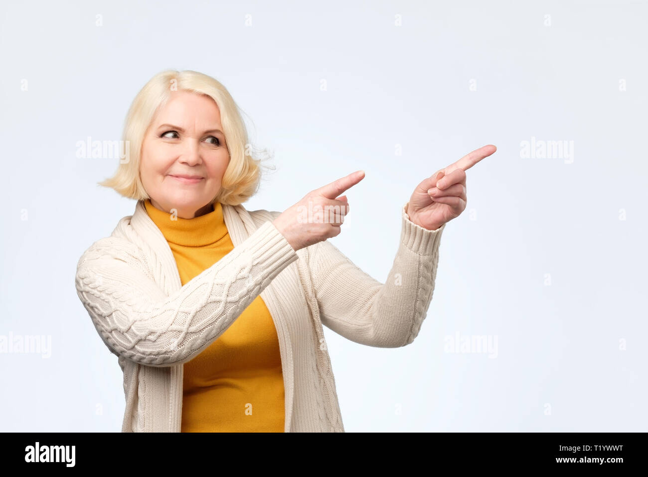 Gerne blonde ältere Frau lächelnd Pointing Finger weg, die etwas Interessantes und Spannendes in Studio Wand Stockfoto