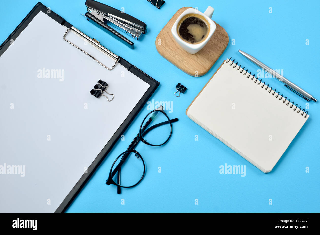 Arbeitsbereich mit leeren Clip Board, Bürobedarf, Kugelschreiber, Notizblock, Brillen, Hefter, Kaffee Tasse und Brillen auf blauem Hintergrund. Flach, zu Stockfoto