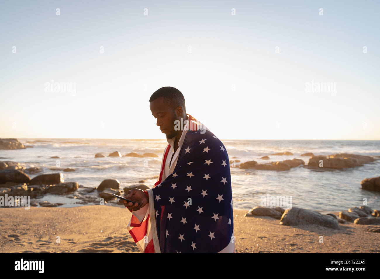 Mann gewickelt amerikanische Flagge bei der Verwendung von Mobile Phone am Strand an einem sonnigen Tag Stockfoto