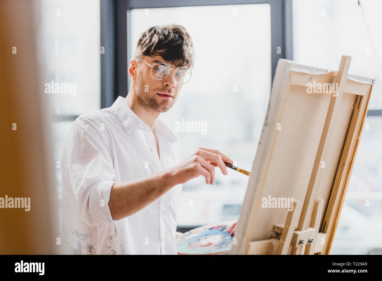 Selektiver Fokus der stattlichen Artist in Gläsern bei Kamera schaut beim Zeichnen auf Leinwand Stockfoto