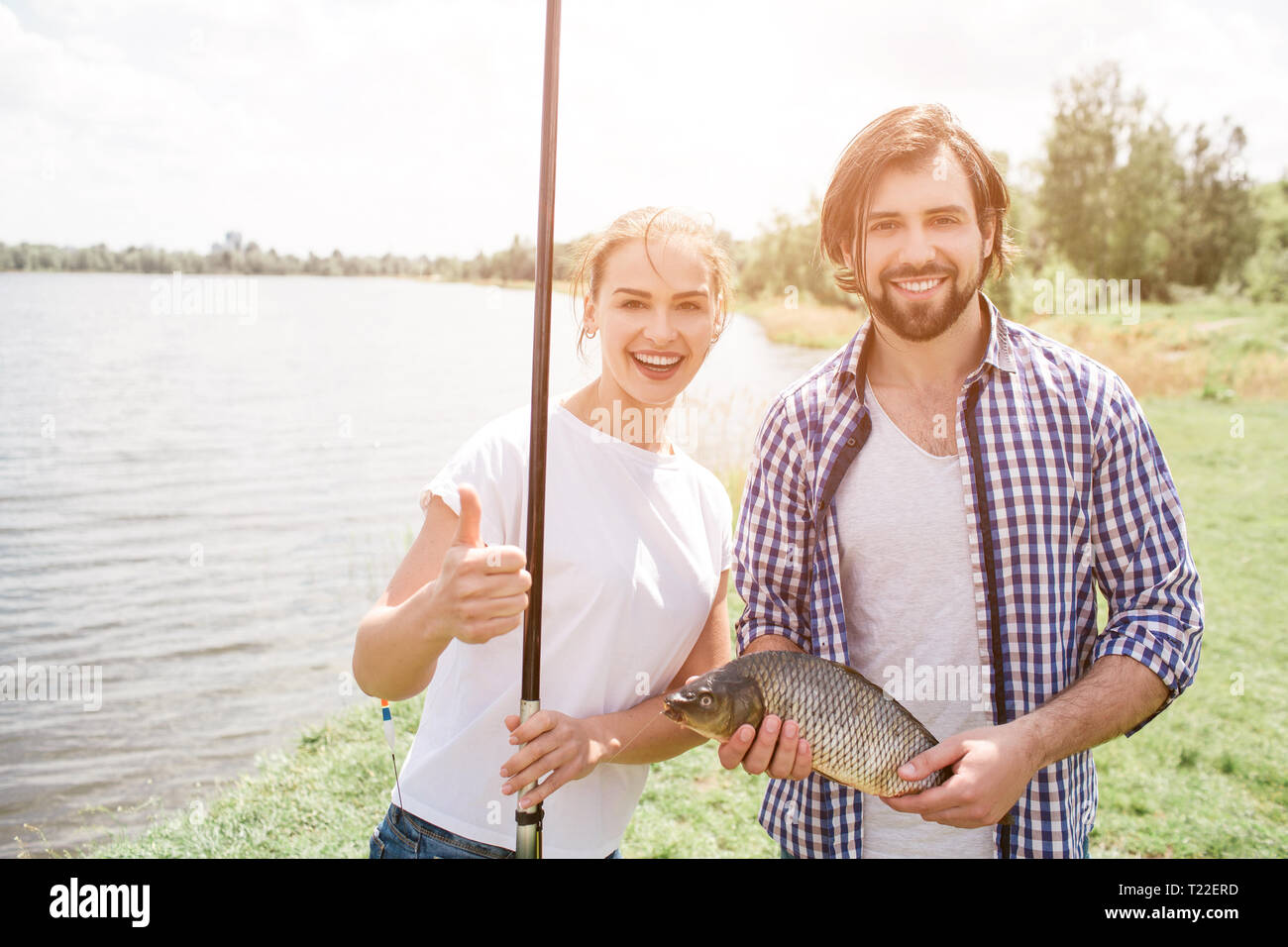 Glückliches Paar steht am Fluss shroe und lächelnd. Sie sucht auf Kamera. Mädchen hält Ihr großes Daumen hoch halten Fisch und-Stange, während der Mensch ist h Stockfoto