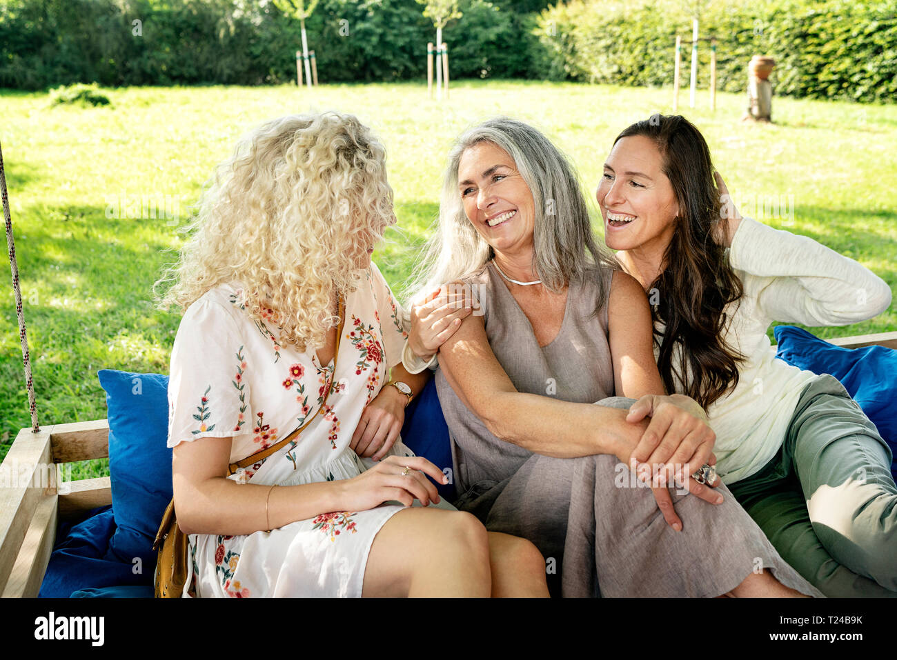 Drei glückliche Frauen verschiedenen Alters sittingon ein hängendes Bett im Garten reden Stockfoto