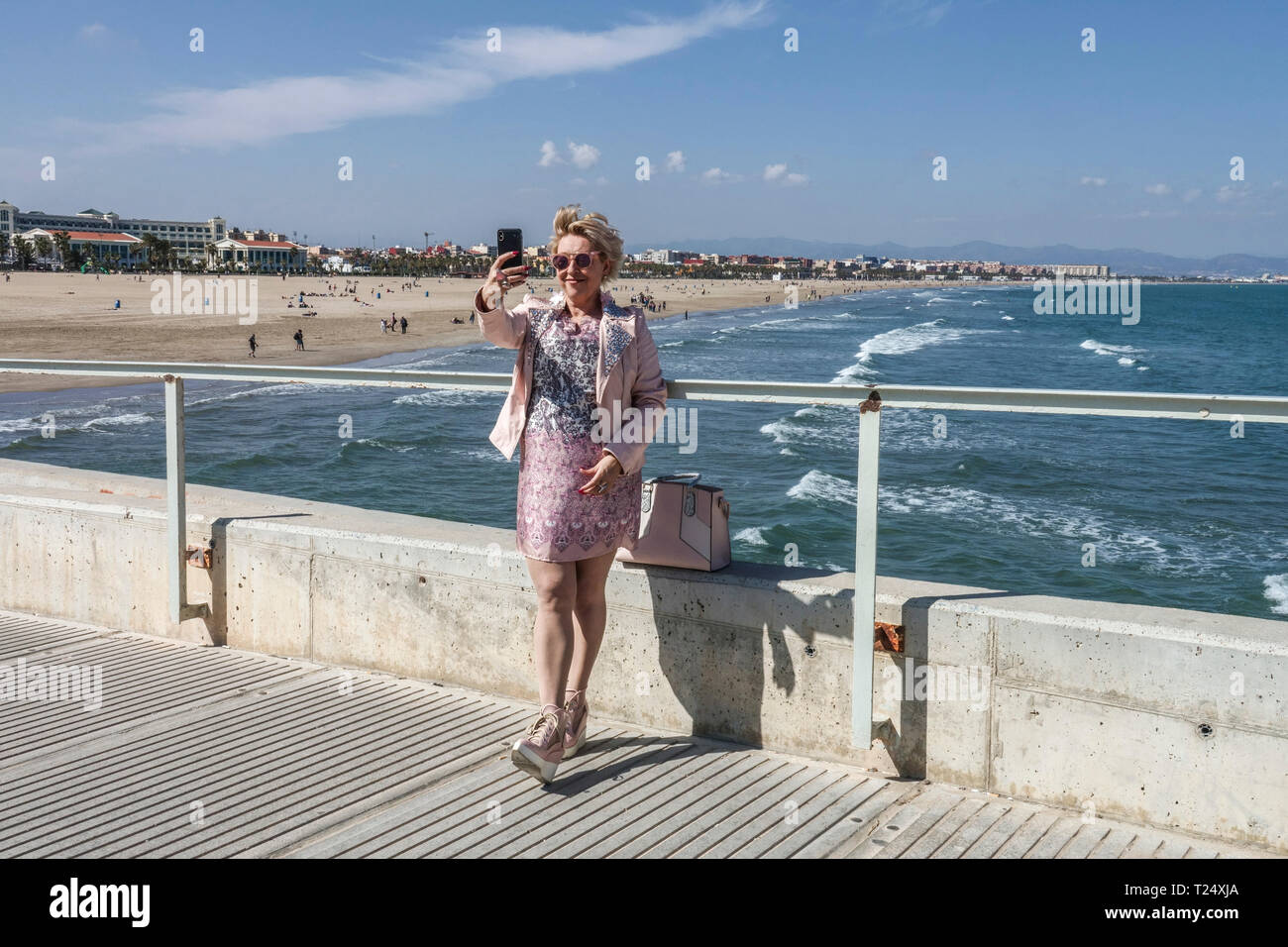 Eine Frau im beigen Anzug, die ein Selfie auf einem Betonpier mit Meer und Malvarrosa Strand nimmt Valencia Meerblick Valencia Spanien Strand Stockfoto