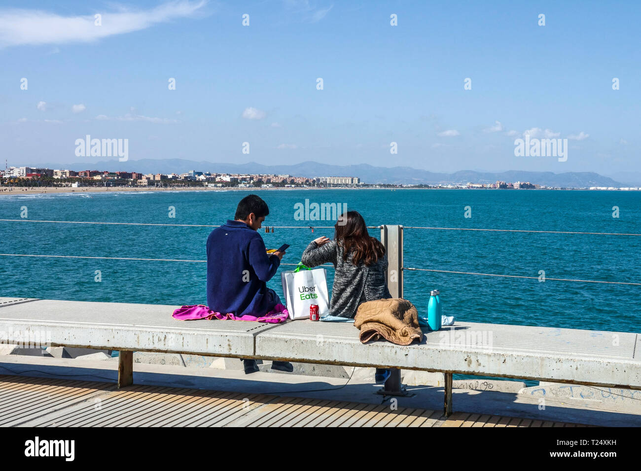 Das Paar machte ein Picknick auf dem Betonpier mit Blick auf das Meer und Malvarrosa Strand das Essen wurde von Uber Eats, Valencia Hafen, Spanien Meerblick geliefert Stockfoto