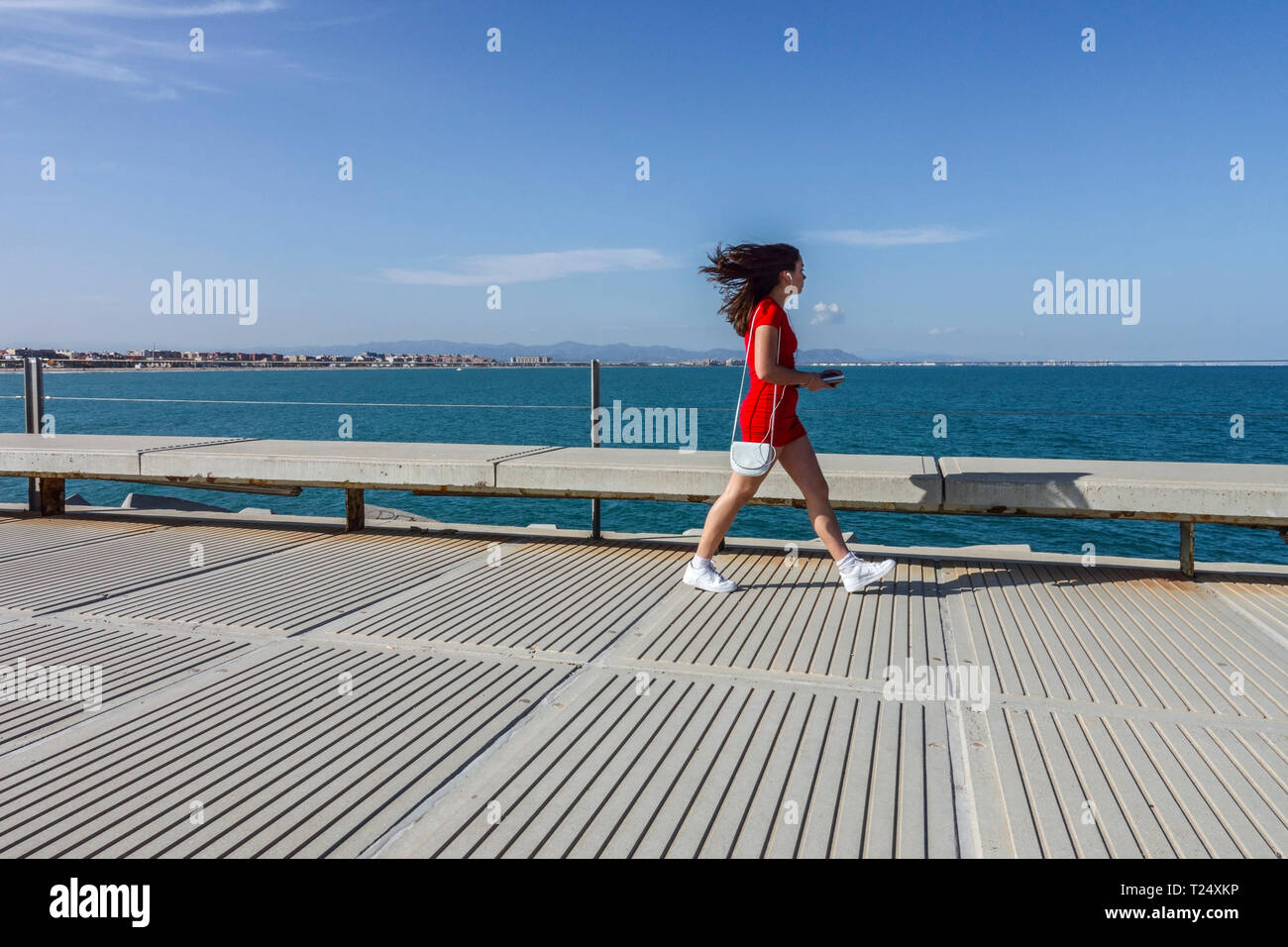 Junge Frau in rotem Kleid, unordentlichen Haaren und Telefon gehen durch den Betonpier, Valencia Hafen, Spanien Frau allein in rotem weiß Stockfoto