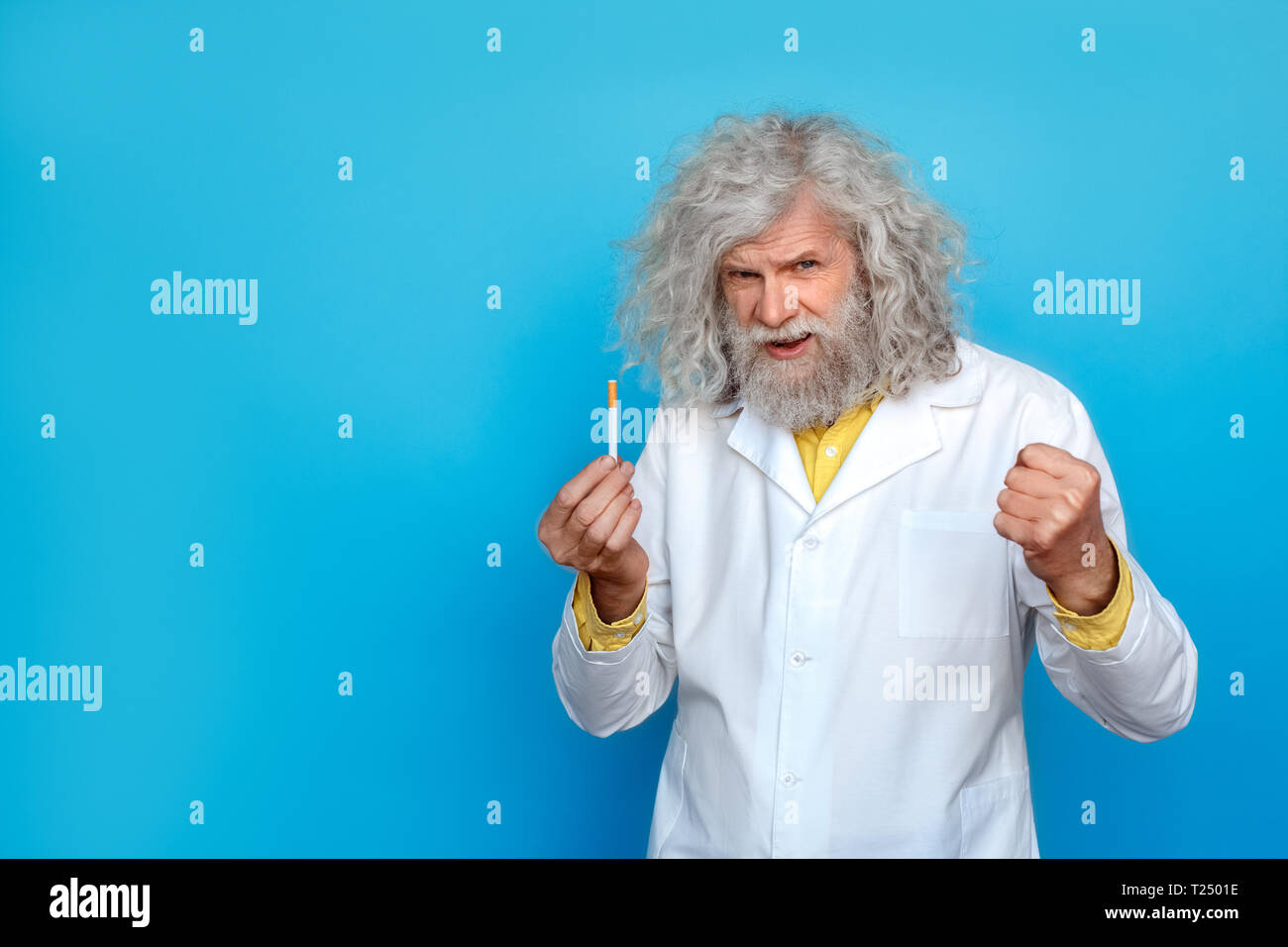 Alte langhaarigen Mann, der Doctor's Kleid Studio auf der blauen Wand holding Zigarette und Hand in Faust suchen Kamera isoliert Sprechen unglücklich Stockfoto