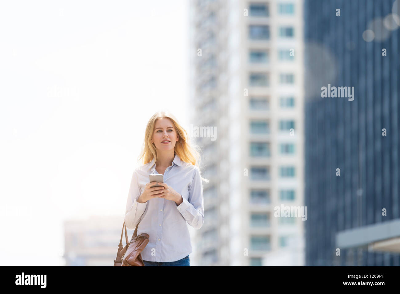 Blonde Frau mit Handtasche und Handy zu Fuß durch die Stadt Stockfoto