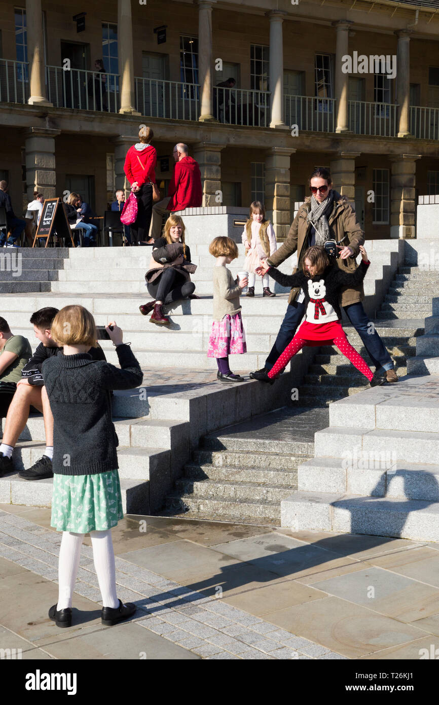 Kind spielt um das Wasserspiel & Besucher/Touristen sitzen und auf den Stufen im Viereck des wiederhergestellten Piece Hall entspannen. Sonnigen Tag/sun. Halifax. England UK. Der Tag war ein außergewöhnlich heißer Tag im Winter. Stockfoto
