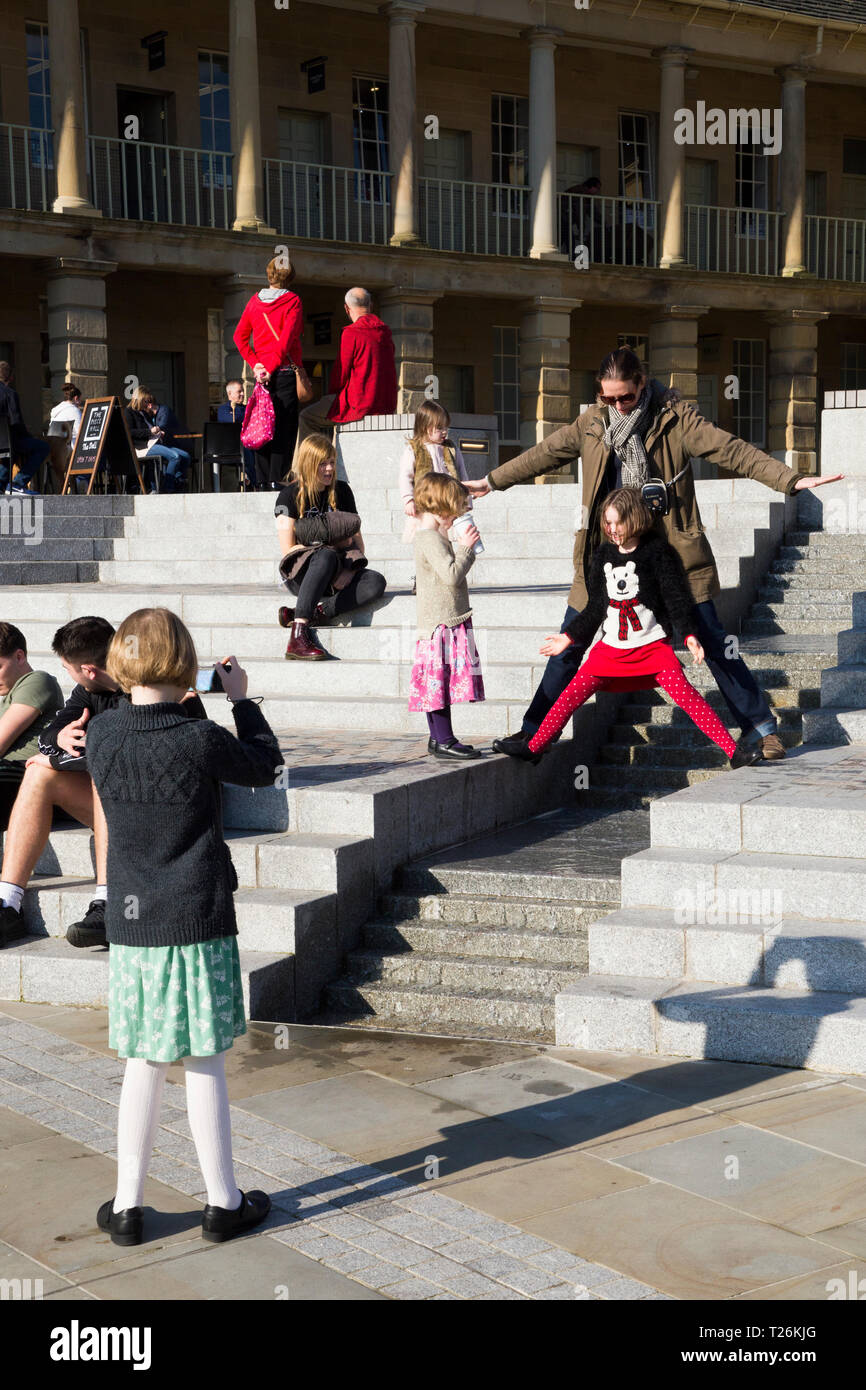 Kind spielt um das Wasserspiel & Besucher/Touristen sitzen und auf den Stufen im Viereck des wiederhergestellten Piece Hall entspannen. Sonnigen Tag/sun. Halifax. England UK. Der Tag war ein außergewöhnlich heißer Tag im Winter. Stockfoto