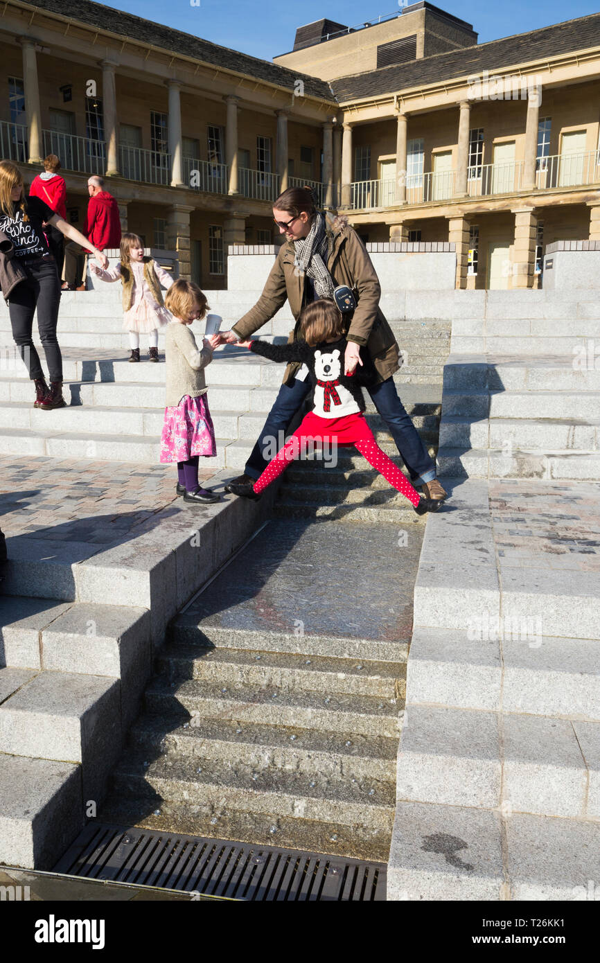 Kind spielt um das Wasserspiel & Besucher/Touristen sitzen und auf den Stufen im Viereck des wiederhergestellten Piece Hall entspannen. Sonnigen Tag/sun. Halifax. England UK. Der Tag war ein außergewöhnlich heißer Tag im Winter. Stockfoto