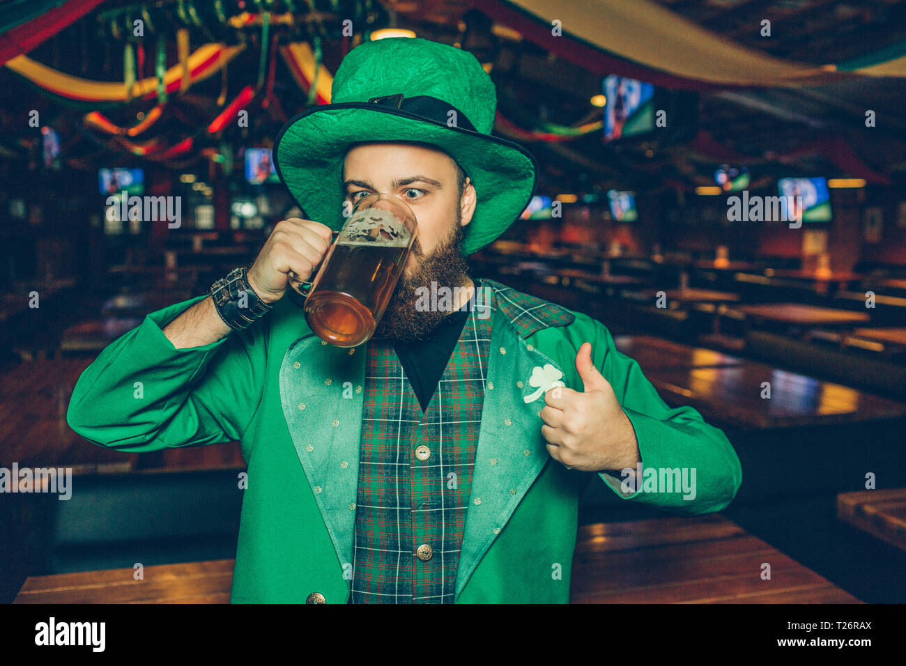 Bild des jungen Mannes im grünen Anzug im Pub Bier trinken aus der Tasse. Er halten grosse Daumen hoch Stockfoto