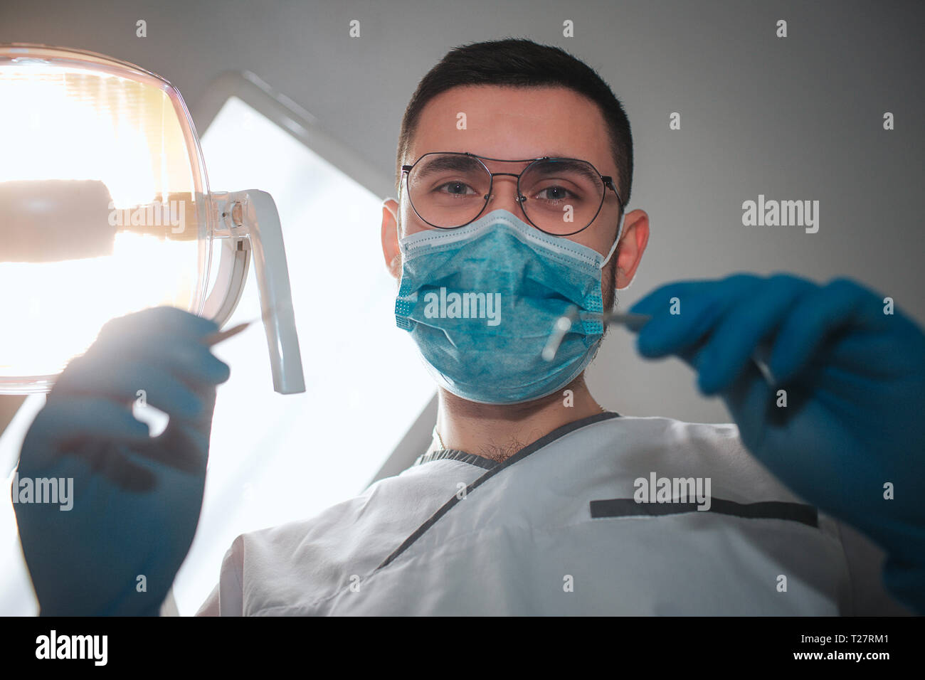 Junge männliche Zahnarzt sehen Sie auf Kamera. Er trägt ein weißes Rob und blaue Maske. Kerl halten Zähne Ausrüstung für Behandlung. Die Lampe eingeschaltet ist. Zahnheilkunde Zimmer. Stockfoto