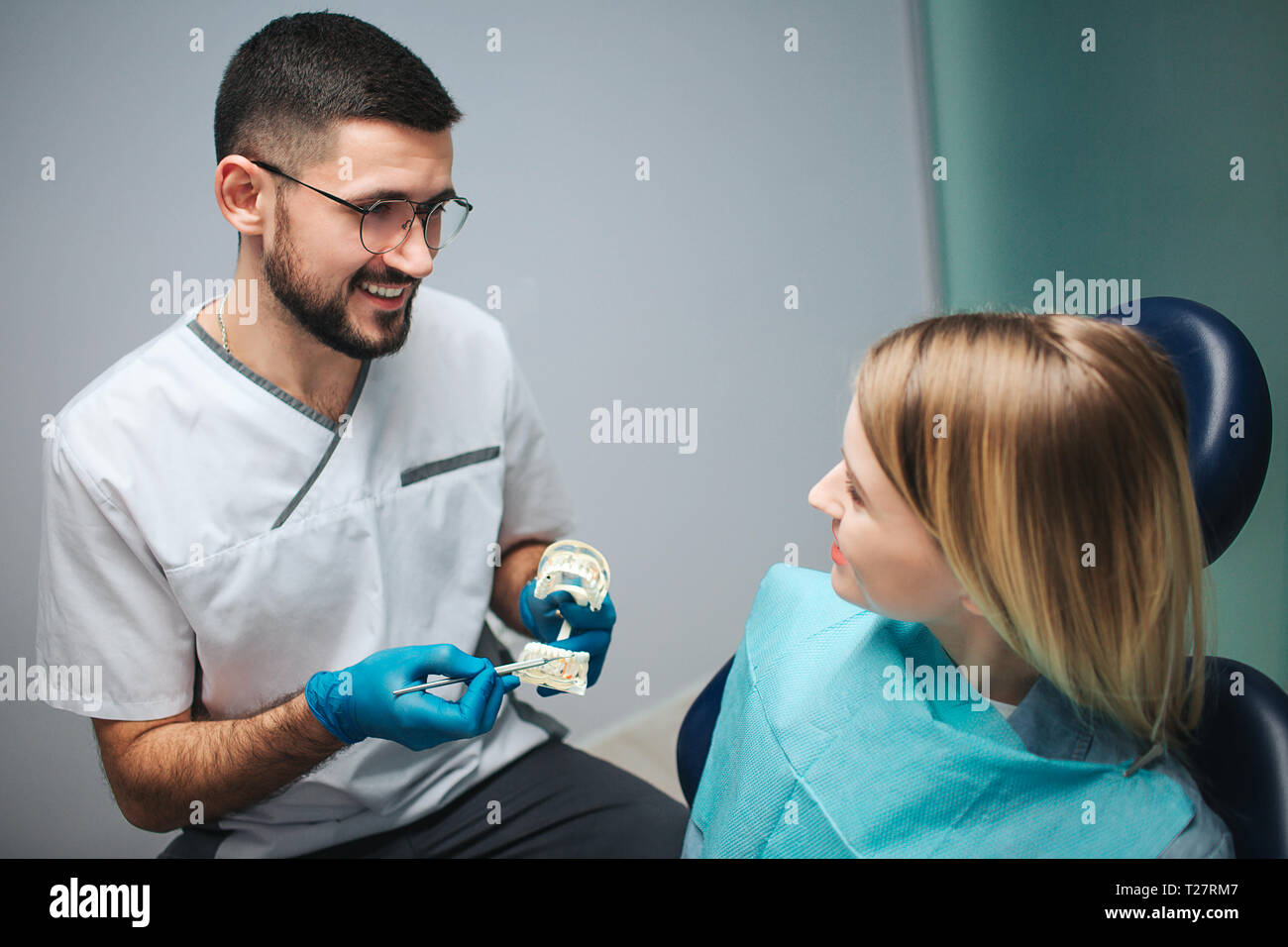 Freundliche nette junge Zahnarzt halten künstliche Kiefer mit Zähnen und Punkt auf Sie in der Zahnheilkunde. Er Blick auf weibliche Client und Lächeln. Junge Frau positiv Stockfoto
